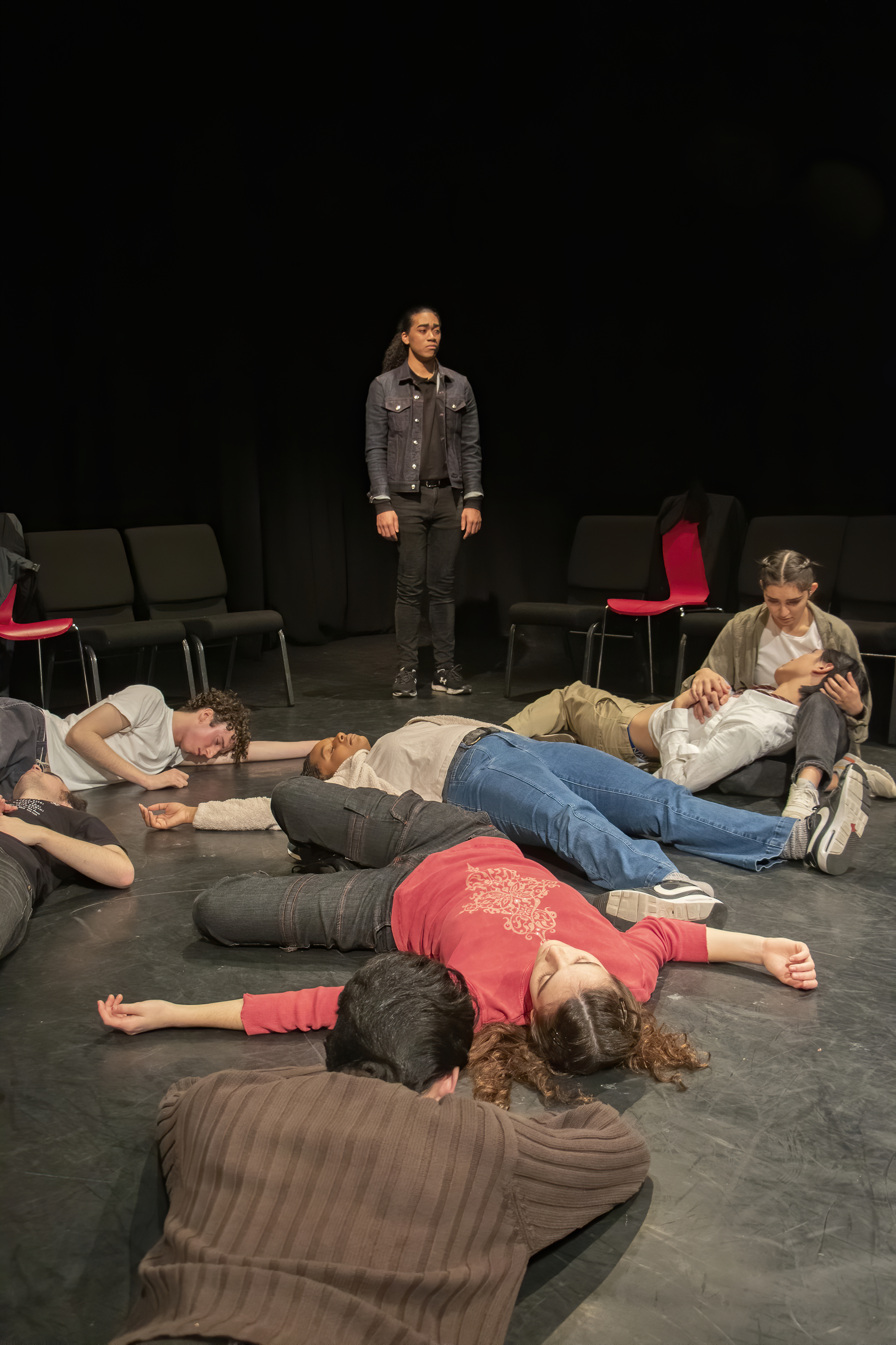 Several students laying on the floor of the theatre as one stands over them, in mid-performance.